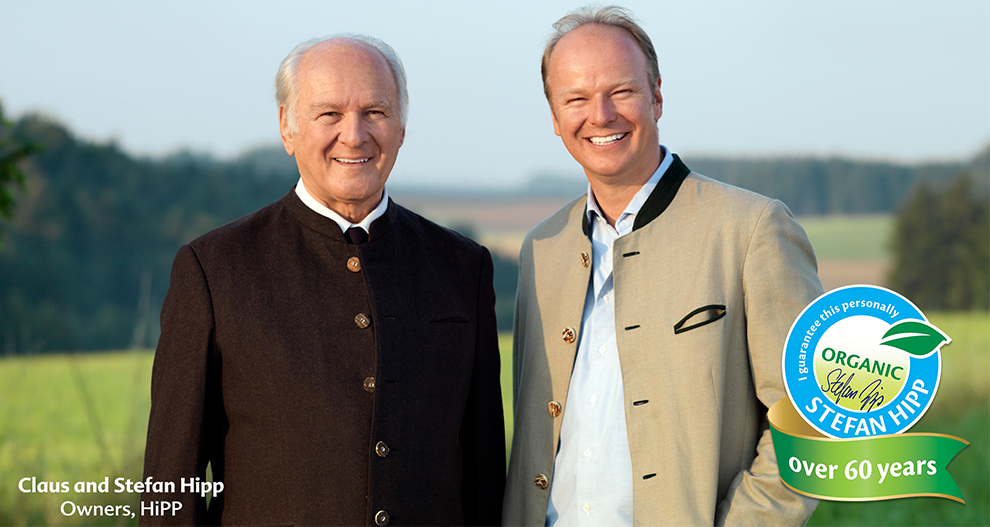 The owners of HiPP, Claus and Stefan Hipp, smiling and posing side by side with a green open field with some trees as the background