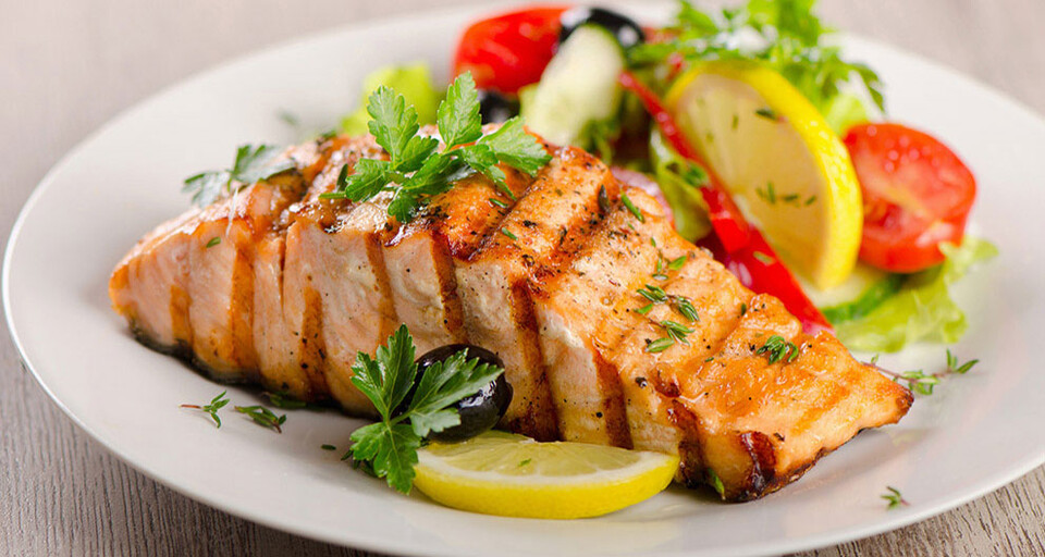 A plate of cooked fish topped with parsley and paired with a side of olives, sliced lemons, sliced tomatoes, and sliced red bell pepper