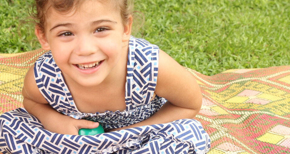 A smiling young girl wearing a picnic dress while seated on a picnic mat on grass