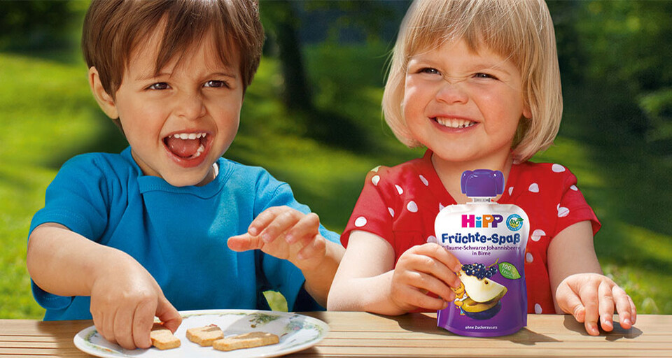 A smiling boy toddler holding a biscuit on a plate while seated beside a girl toddler holding a HiPP food product