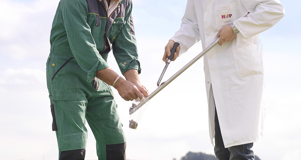 A farmer in an modern agricultural jumpsuit and a HiPP employee in a white laboratory coat with a HiPP logo collecting samples from the field