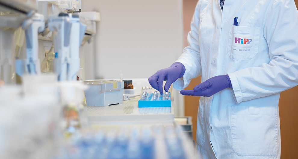 A HiPP employee in a laboratory environment wearing a white laboratory coat with the HiPP brand logo embedded on the chest pocket handling laboratory samples